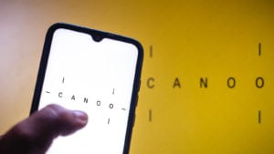 Canoo (GOEV) logo displayed on smartphone screen as well as in background on yellow wall