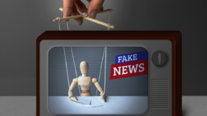 A concept image of a TV displaying a mannequin sitting at a desk with the words "fake news" in a news broadcast-style format with a hand holding marionette strings over the mannequin.