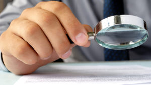 A photo of a hand holding a magnifying glass and looking at a paper.