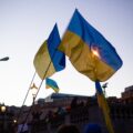 Two Ukrainian flags fly in the wind, with a crowd below them and buildings in the background