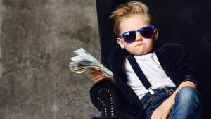 A photo of a young boy wearing sunglasses, jeans, a blazer, a white shirt and suspenders holding money in various denominations in one hand and sitting in a plush chair.