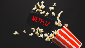 An image of a phone with the Netflix logo on the screen, laying next to a container of popcorn with popcorn splayed across