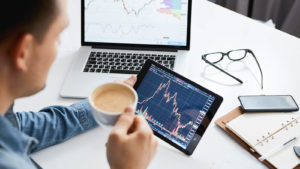 a person drinks coffee while looking at stock charts on their tablet. a laptop, glasses, and notebook are on the desk in front of them.